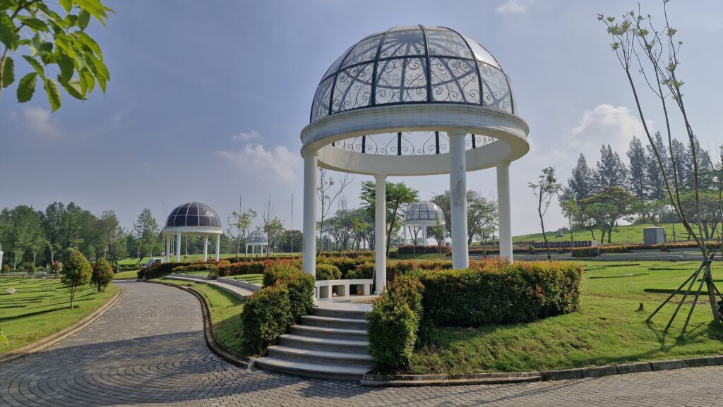 Paviliun megah bergaya klasik di area taman pemakaman San Diego Hills dikelilingi tanaman hijau dan jalan setapak berbatu, menciptakan suasana tenang dan elegan.